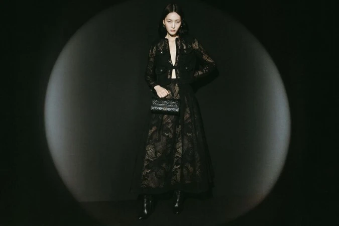 Elegant woman in black lace jacket and skirt holding a purse, posing in a dark photo studio with spotlight. Fashion photography.