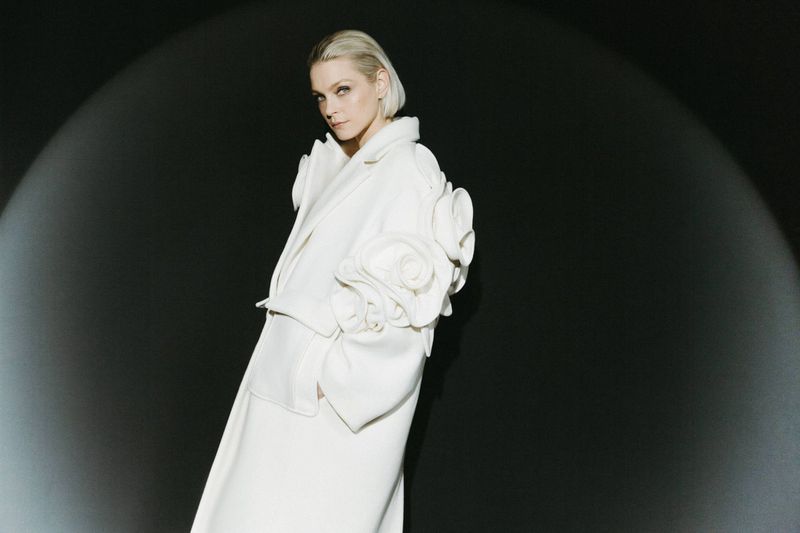 Woman in cream-colored coat with rosette design on sleeve, spotlight on face, against a dark background in a fashion forward pose.