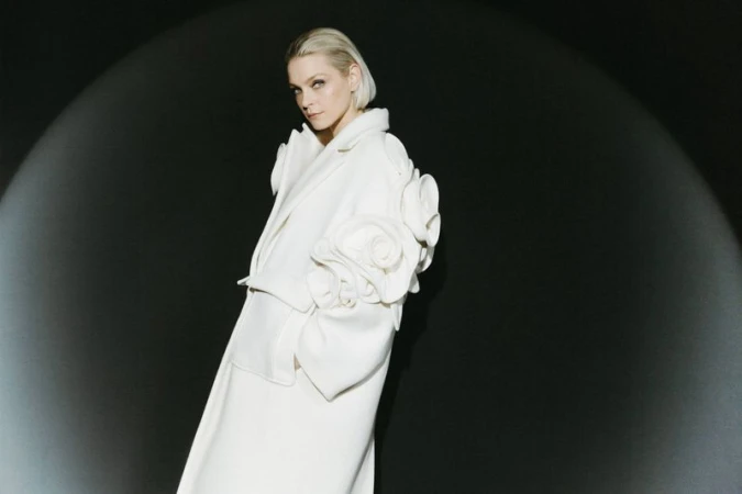 Woman in cream-colored coat with rosette design on sleeve, spotlight on face, against a dark background in a fashion forward pose.