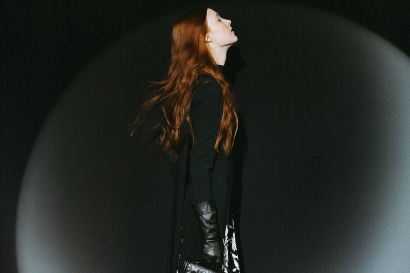 Profile view of a woman with long red hair, wearing a black dress and gloves, standing in front of a dark background illuminated by spotlights.