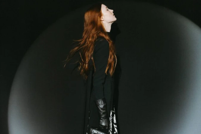 Profile view of a woman with long red hair, wearing a black dress and gloves, standing in front of a dark background illuminated by spotlights.