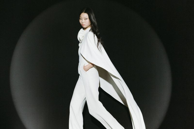 An Asian fashion model in white cape and pants poses in front of a black background lit by a spotlight during a runway show.