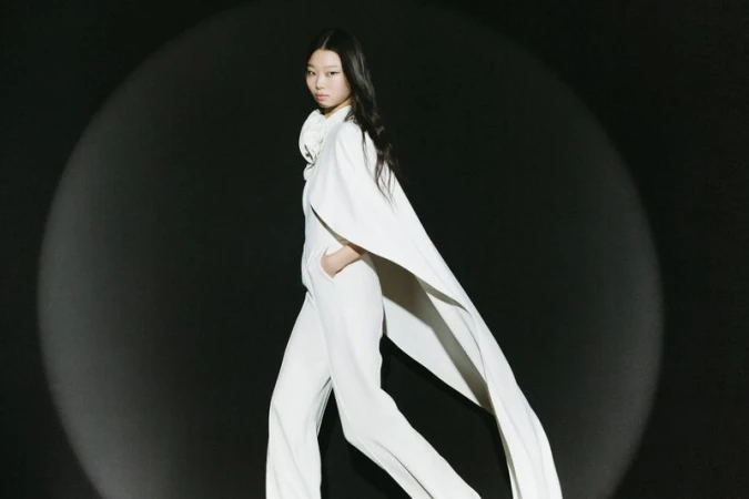 An Asian fashion model in white cape and pants poses in front of a black background lit by a spotlight during a runway show.