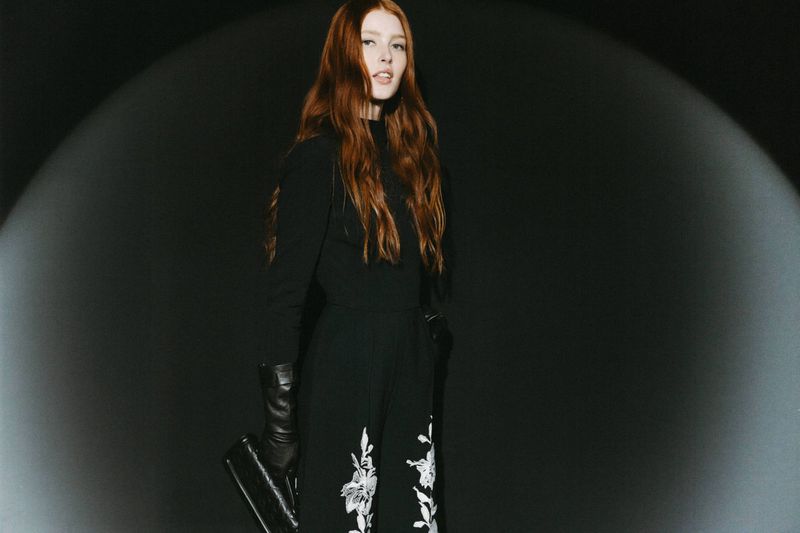 Elegant woman with red hair poses in front of a dark background with spot lighting, wearing black attire and holding a handbag.