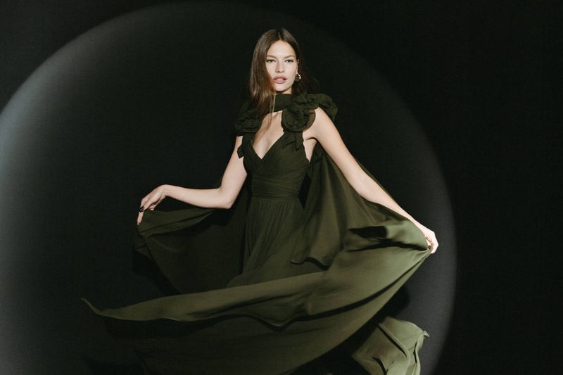A woman in a flowing green gown poses against a dark backdrop with soft lighting, showcasing an elegant and timeless fashion aesthetic.