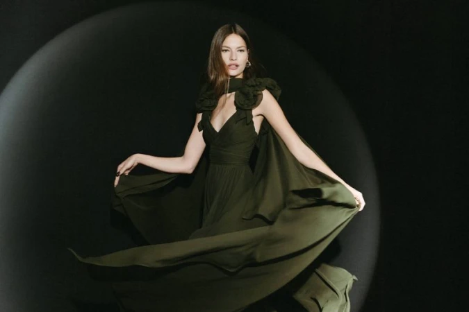A woman in a flowing green gown poses against a dark backdrop with soft lighting, showcasing an elegant and timeless fashion aesthetic.