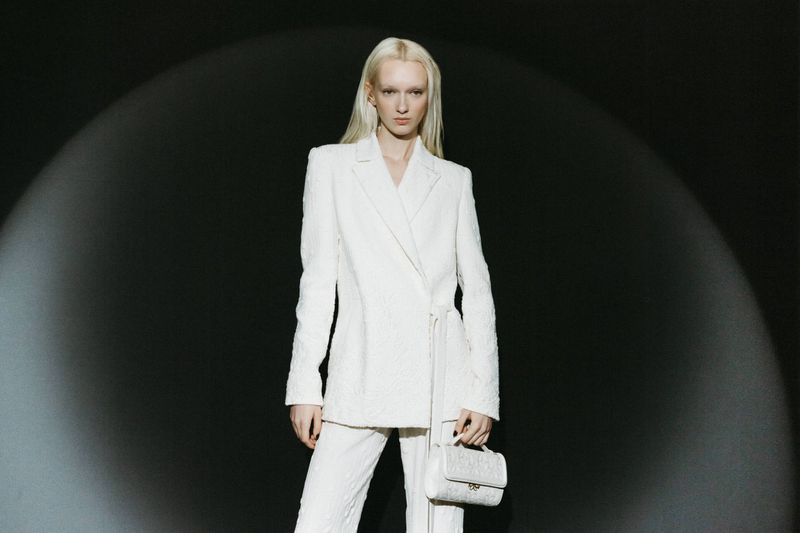 A woman with pale skin and long blonde hair poses in a bright white suit and pants with a matching small handbag against a dark background.