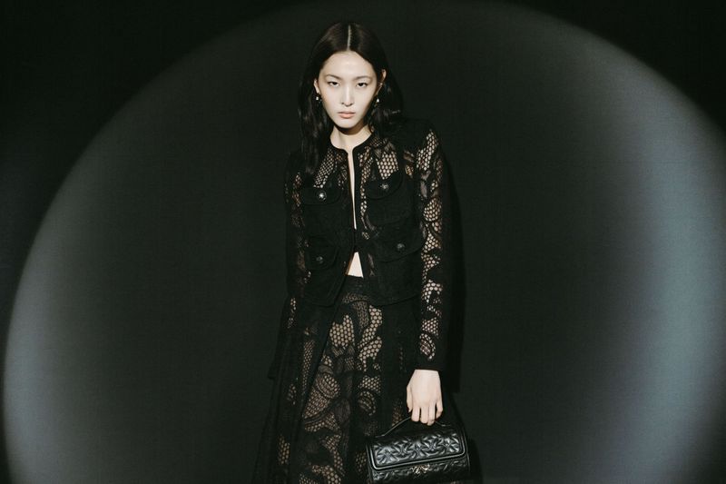 Woman in a black lace jacket and skirt holding a quilted handbag, posing in a studio with a spotlight effect on a dark background.