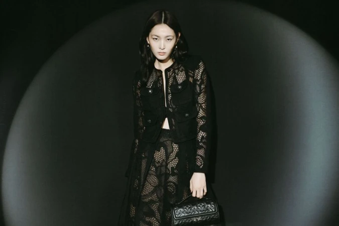 Woman in a black lace jacket and skirt holding a quilted handbag, posing in a studio with a spotlight effect on a dark background.