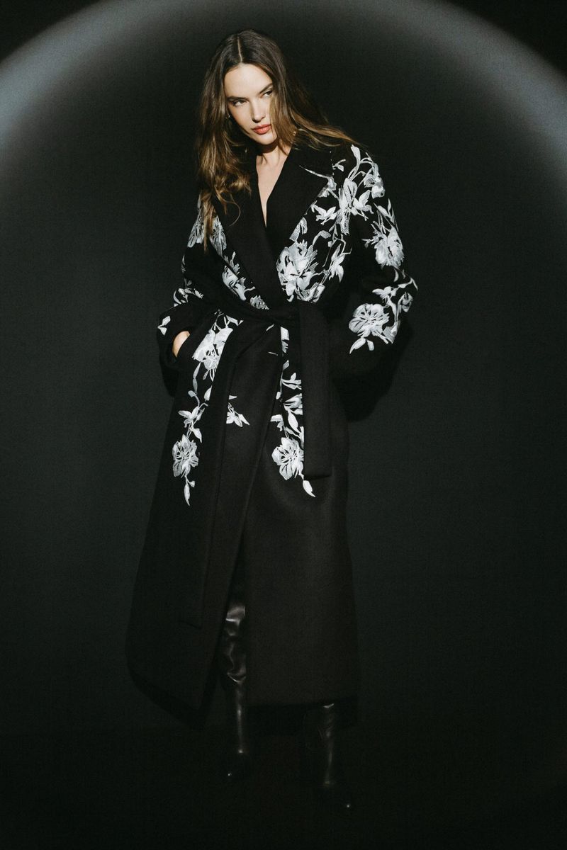 Elegant adult female posing in a black belted coat with white floral embroidery against a dark backdrop in a studio setting.