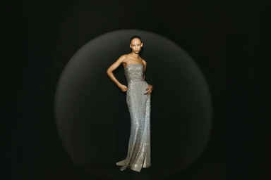 Model posing in a strapless shimmering silver sequin evening gown illuminated inside a spotlight against a black background.