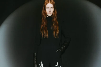 Model with long red hair wearing a black turtleneck and black leather gloves standing under a spotlight