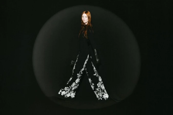 Redhead model walking in wide-leg black trousers with white floral embroidery inside a spotlight against a black background.
