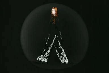 Redhead model walking in wide-leg black trousers with white floral embroidery inside a spotlight against a black background.