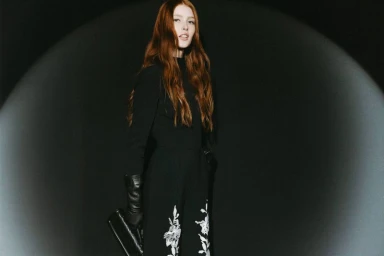 Model with long auburn hair wearing a black embroidered jumpsuit and leather gloves in a spotlight against a dark backdrop.