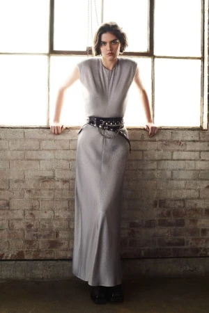 A woman stands in front of a window wearing a grey maxi dress with a studded belt in an indoor setting with brick walls.