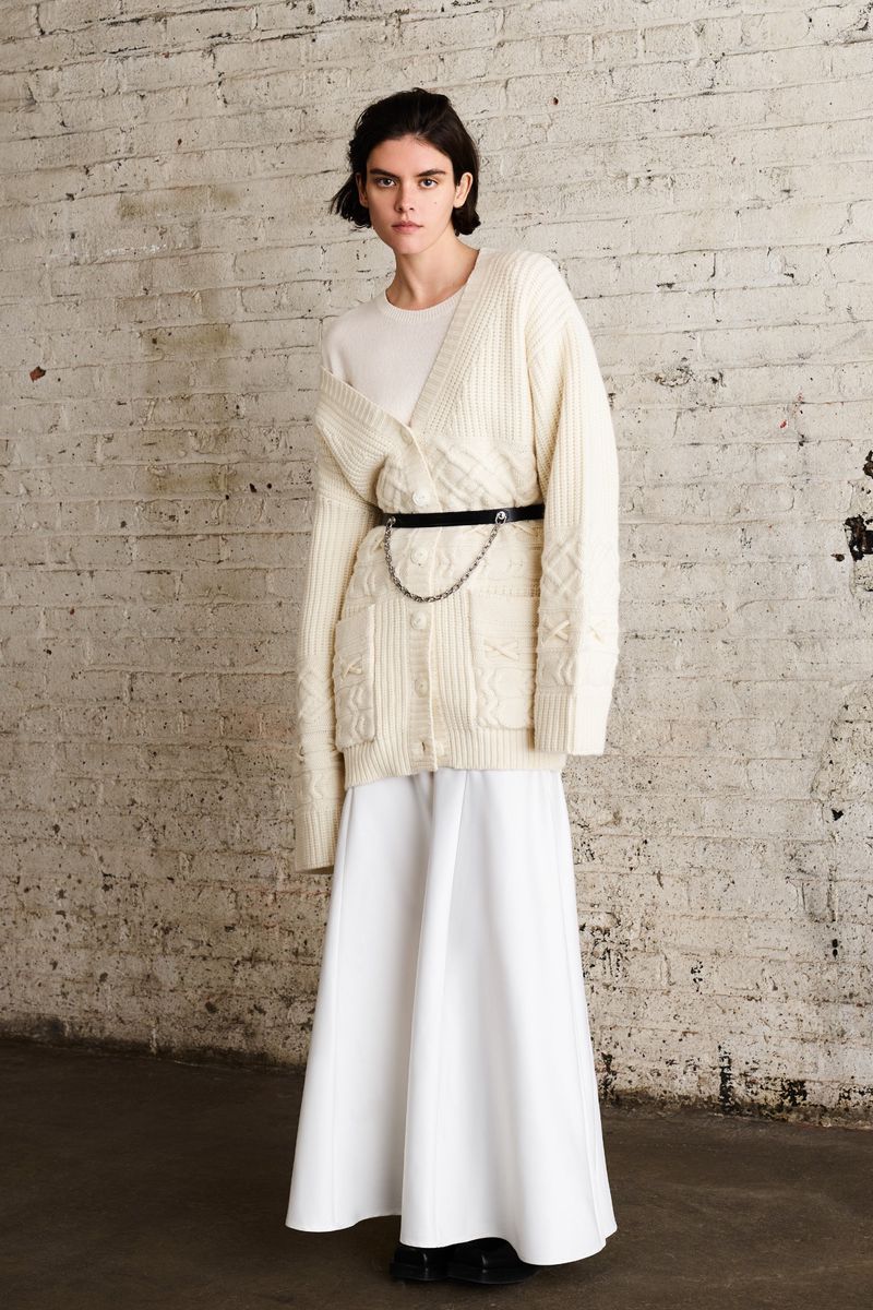 A woman stands in front of a textured wall, wearing a cream-colored cardigan, matching top, white skirt, and a belt with a silver chain.
