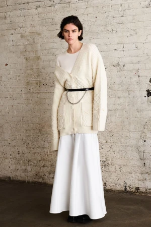 A woman stands in front of a textured wall, wearing a cream-colored cardigan, matching top, white skirt, and a belt with a silver chain.