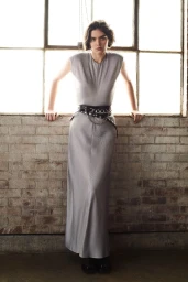 Model in grey maxi dress with layered black belts leaning against a brick wall in front of windows.