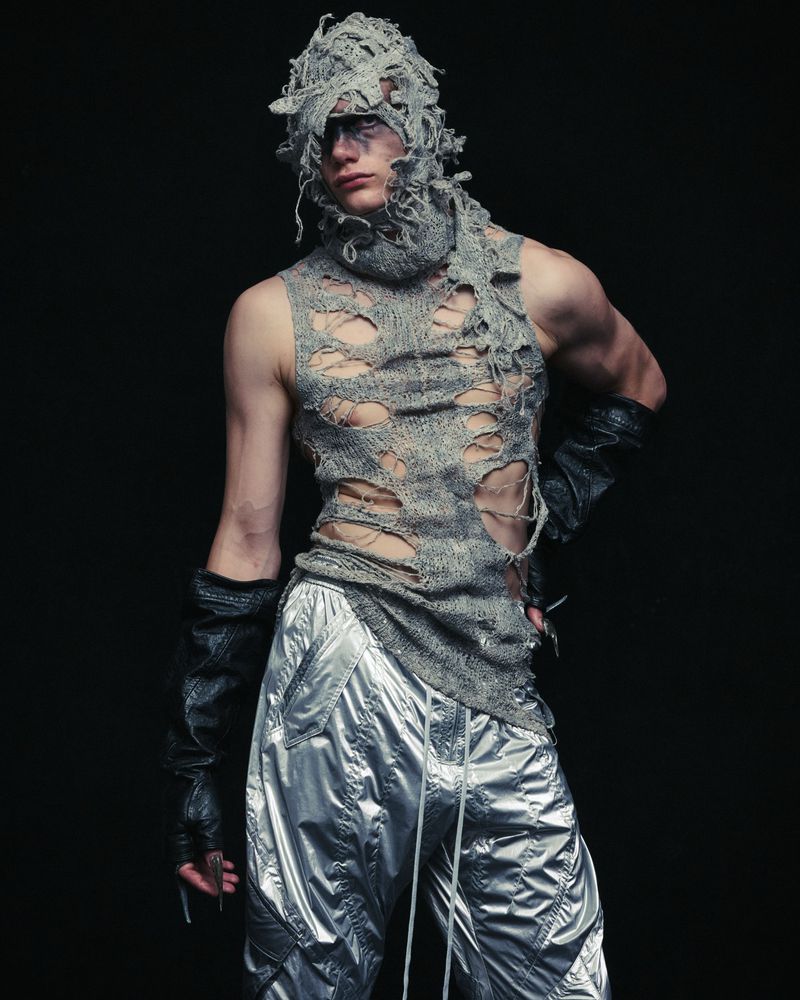 Adult male fashion model poses in ripped gray knitted top and shiny silver pants in studio against dark backdrop with dramatic lighting.