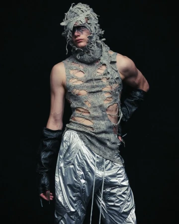 Adult male fashion model poses in ripped gray knitted top and shiny silver pants in studio against dark backdrop with dramatic lighting.