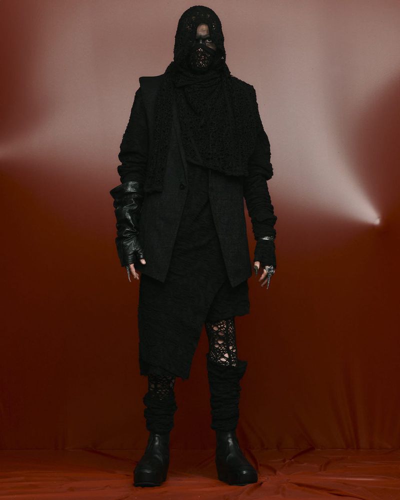 A person in an all-black outfit with a textured jacket, hooded mask, and fingerless gloves stands against a gradient red background.