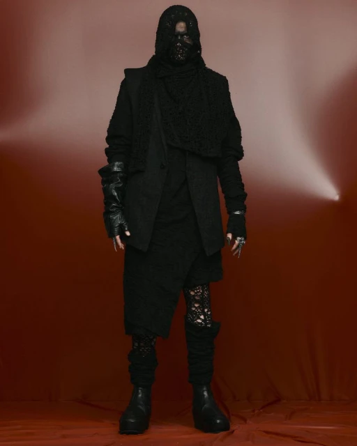A person in an all-black outfit with a textured jacket, hooded mask, and fingerless gloves stands against a gradient red background.