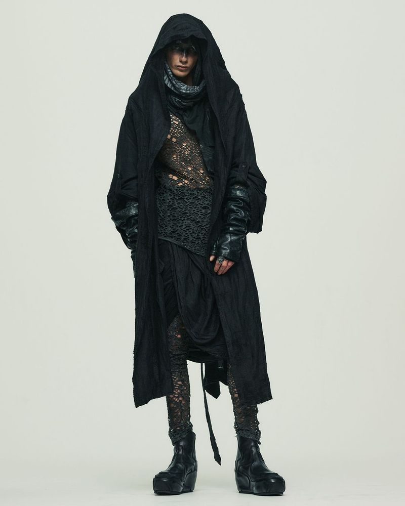 Full-length portrait of a model in dark, avant-garde fashion, featuring draped layers, a hood, and textured fabrics against a plain backdrop.