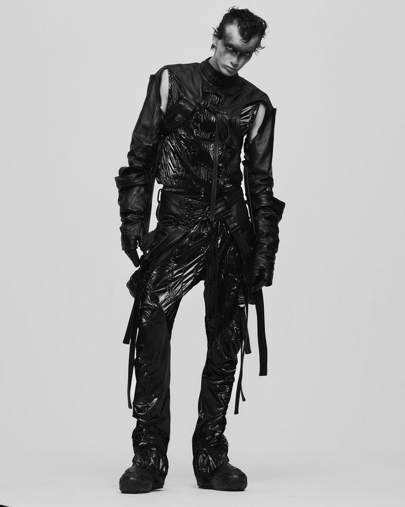 Monochrome full shot of a person posing in a futuristic-style black outfit, against a light grey backdrop, showcasing avant-garde fashion.