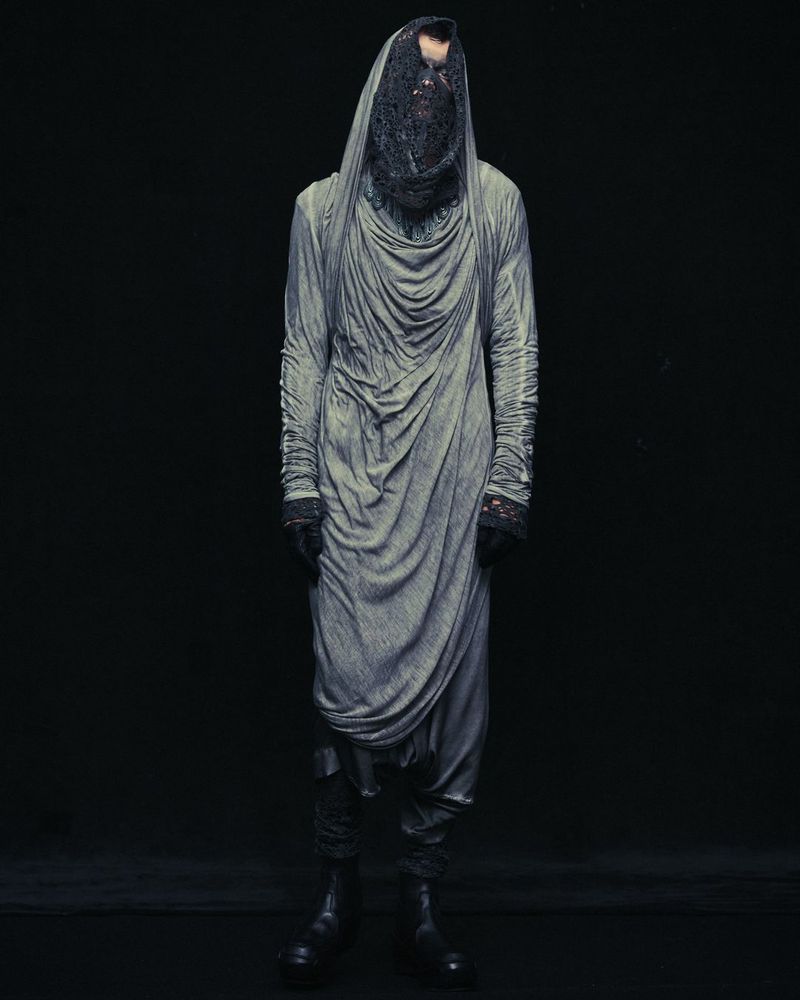 Full-length studio portrait of a person wearing a draped grey robe with a dark mesh covering their head and face, against a black background.