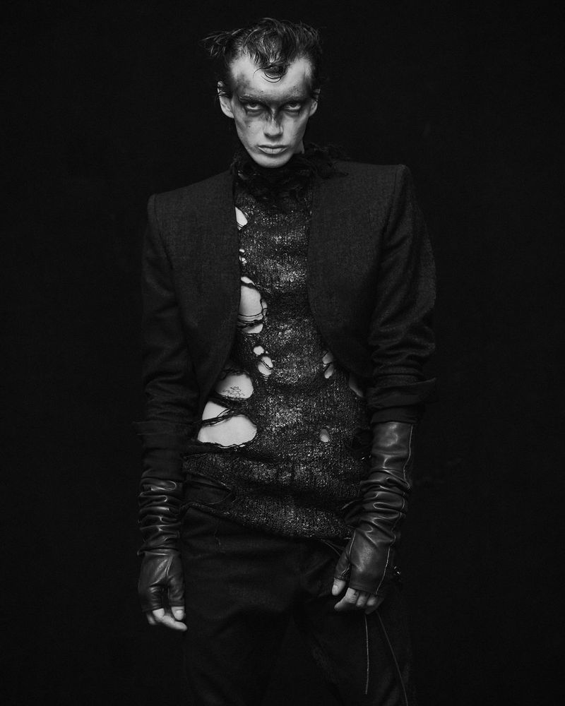 Black and white portrait of a person in a torn black shirt, dark eye makeup, and gloves, posing in front of a solid black background.