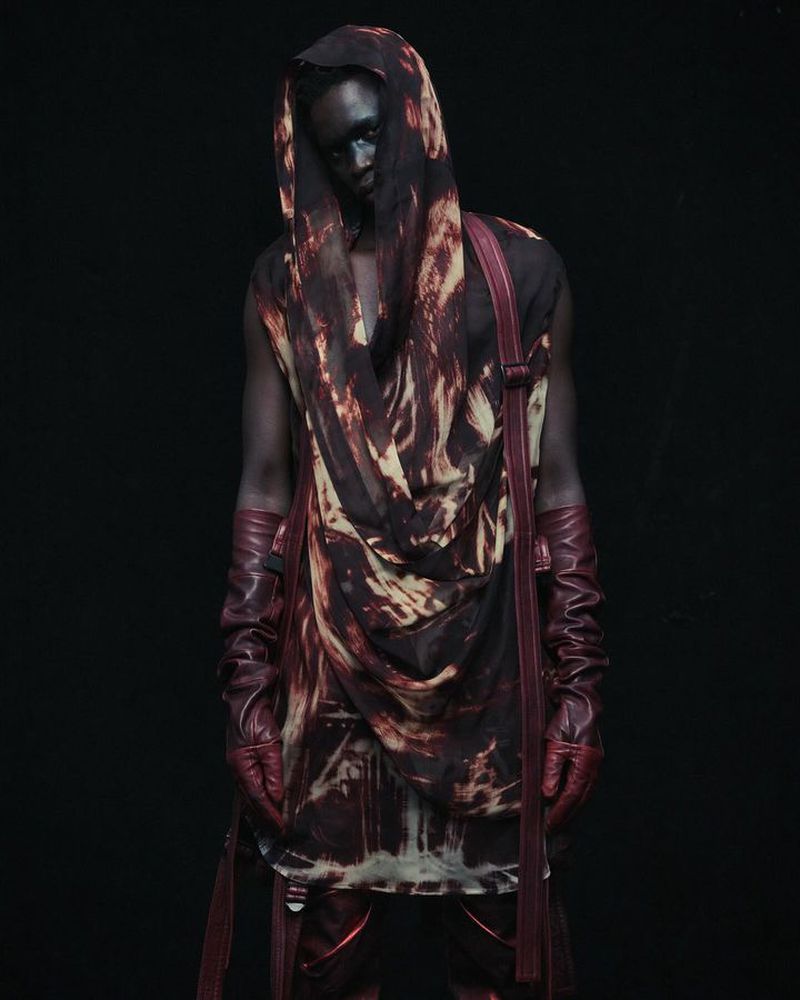 Fashionable person in an abstract-printed hooded top with matching gloves poses in front of a dark background in a studio shot.