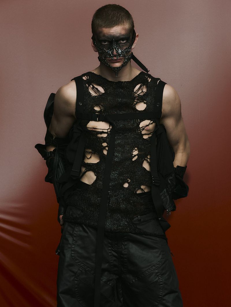Adult male wearing a black distressed top and black mask poses against a gradient red backdrop in a studio portrait. Dark, edgy fashion.