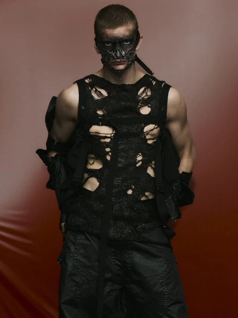 Adult male wearing a black distressed top and black mask poses against a gradient red backdrop in a studio portrait. Dark, edgy fashion.