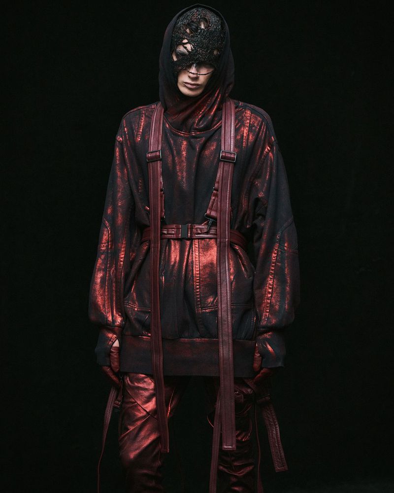 Androgynous person in hooded metallic red-and-black outfit and textured mask against dark backdrop in a fashion photography style.