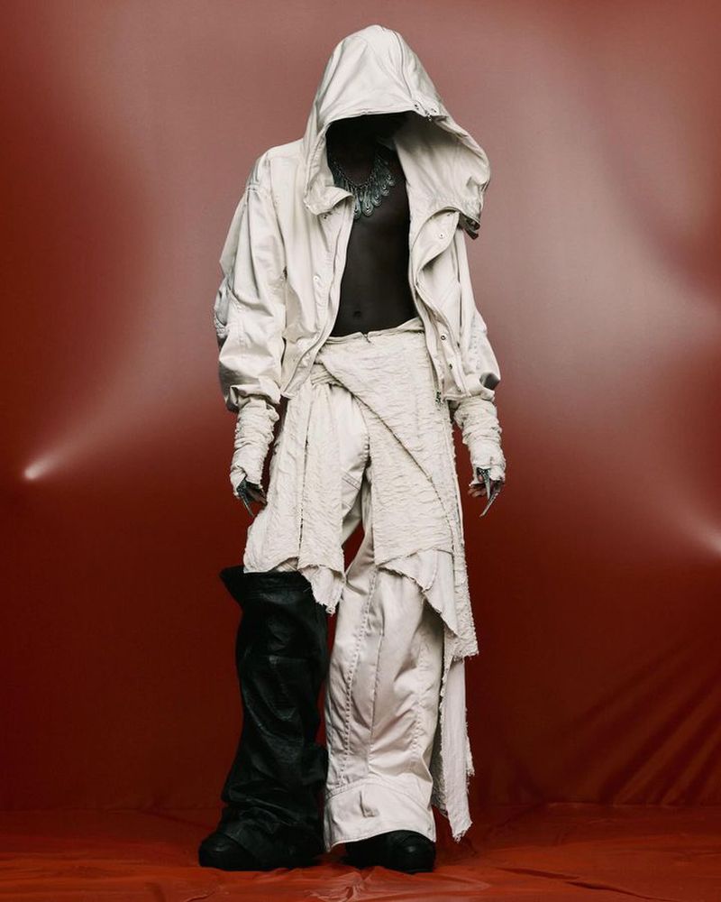 Fashion shot of a person wearing a white hooded jacket and pants with a draped cloth, with one black leather boot, on a red background.
