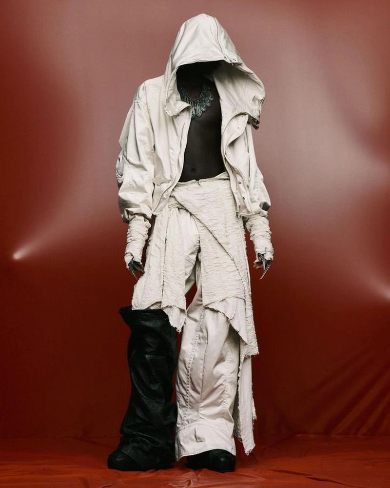 Model wearing an oversized off-white hooded jacket and layered wrap trousers against a terracotta studio backdrop.