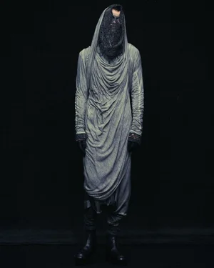 Model wearing a draped light grey hooded tunic and dark face veil against a black background for Julius S/S 25 lookbook.