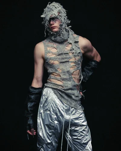 Model wearing a distressed shredded grey knit balaclava top and metallic silver pants for Julius against a black background.