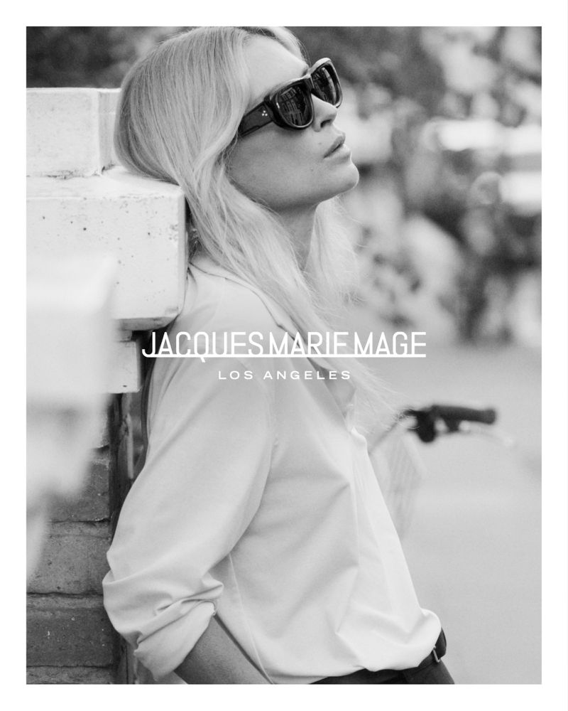 Erin Wasson wearing bold dark sunglasses and a white collared shirt leaning against a pillar in a black and white portrait.