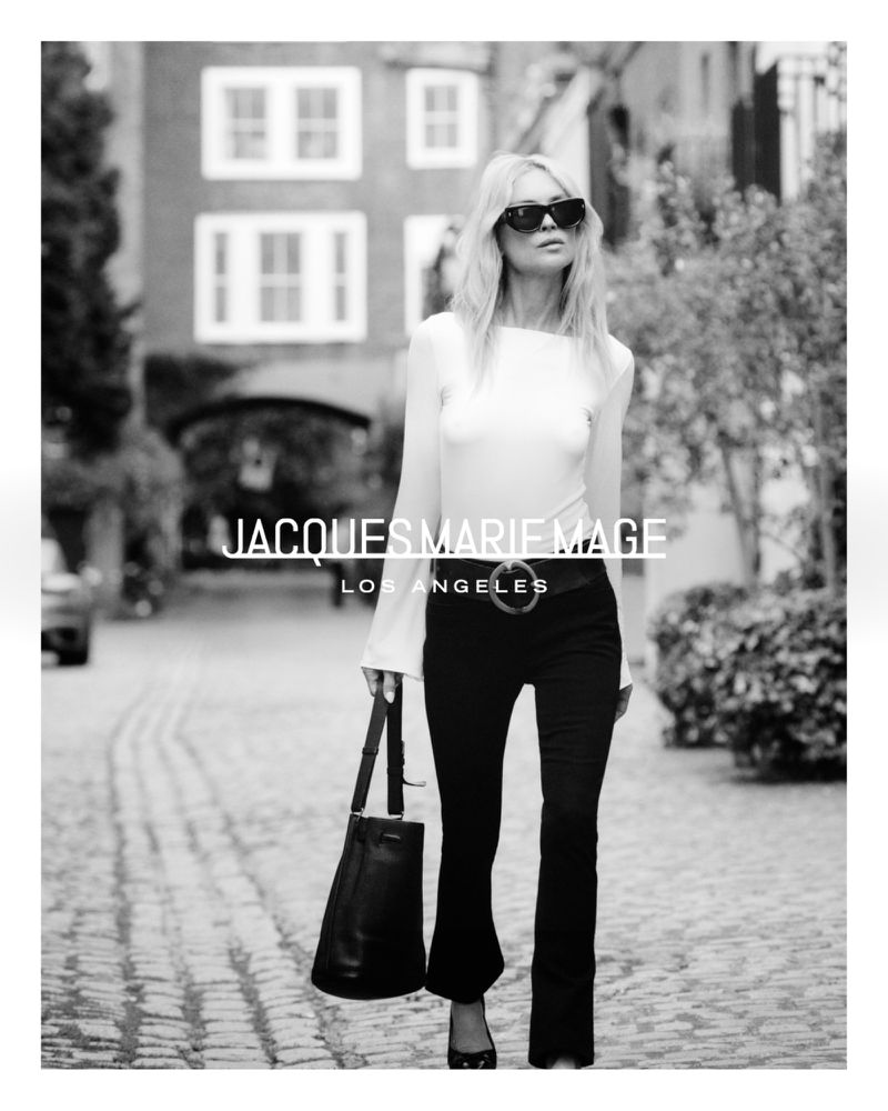 Blonde model in sunglasses and black pants walks on a cobblestone path holding a leather bag in a black and white fashion shot.