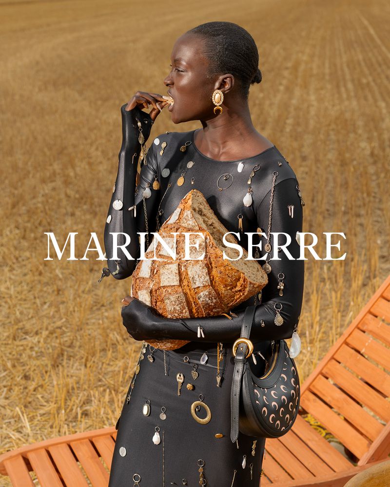 A Black woman in a black outfit holds bread in a wheat field, wearing gold earrings and a purse, looking to the side with text overlay.