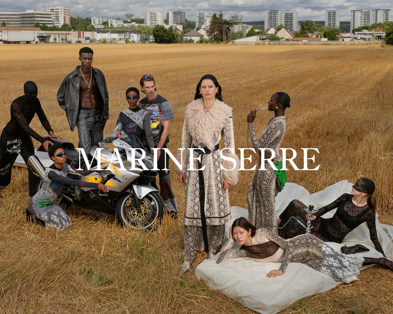 Editorial fashion shot with a diverse group of models in wheat field; cityscape visible in the background; MARINE SERRE logo overlay.
