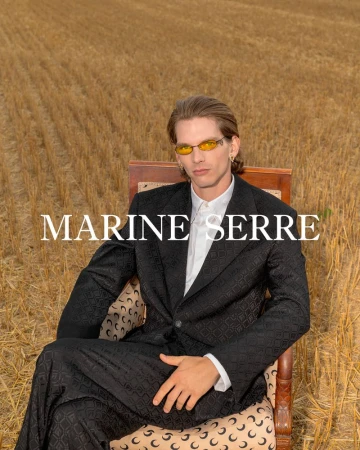 An adult male wearing sunglasses, a white shirt, and patterned suit, seated in a chair in a field of golden wheat, fashion-editorial style.