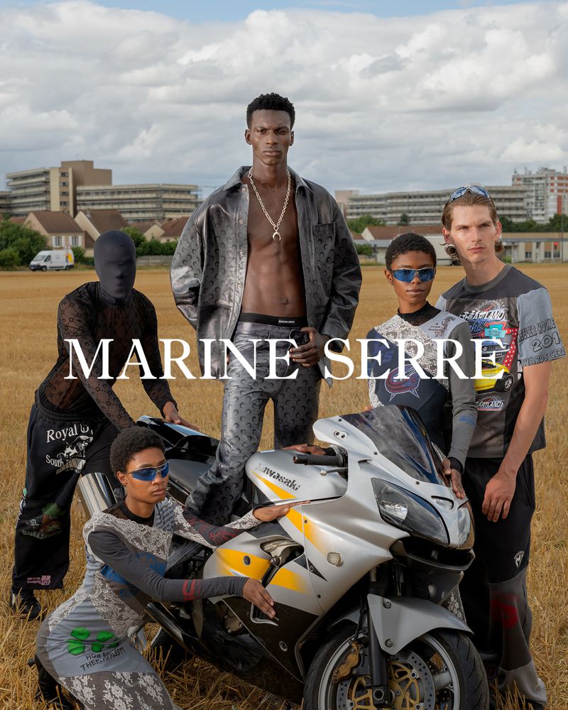 Fashion editorial featuring a group of models posing with a motorcycle in a field under a cloudy sky, wearing designer clothing. 
