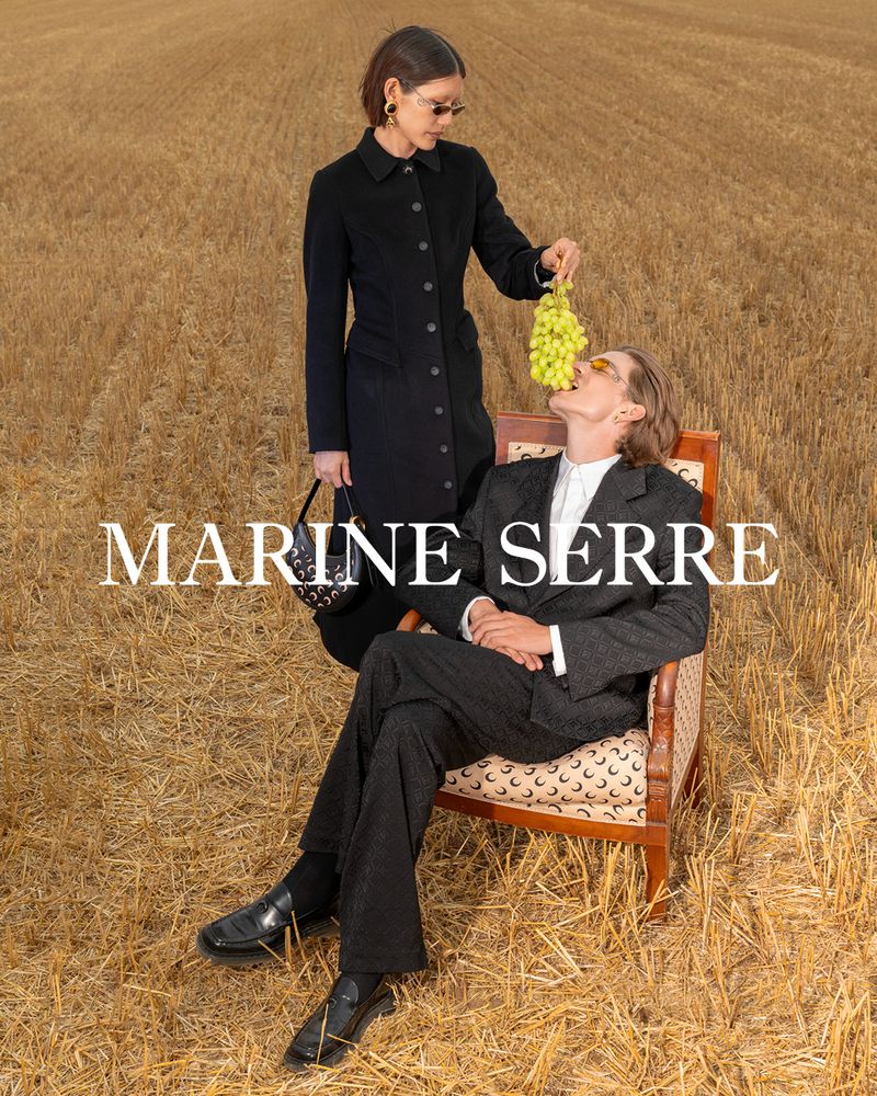 Two fashionably dressed people pose in a wheat field; one feeds the other green grapes in a sun-drenched outdoor shot with Marine Serre text overlay.