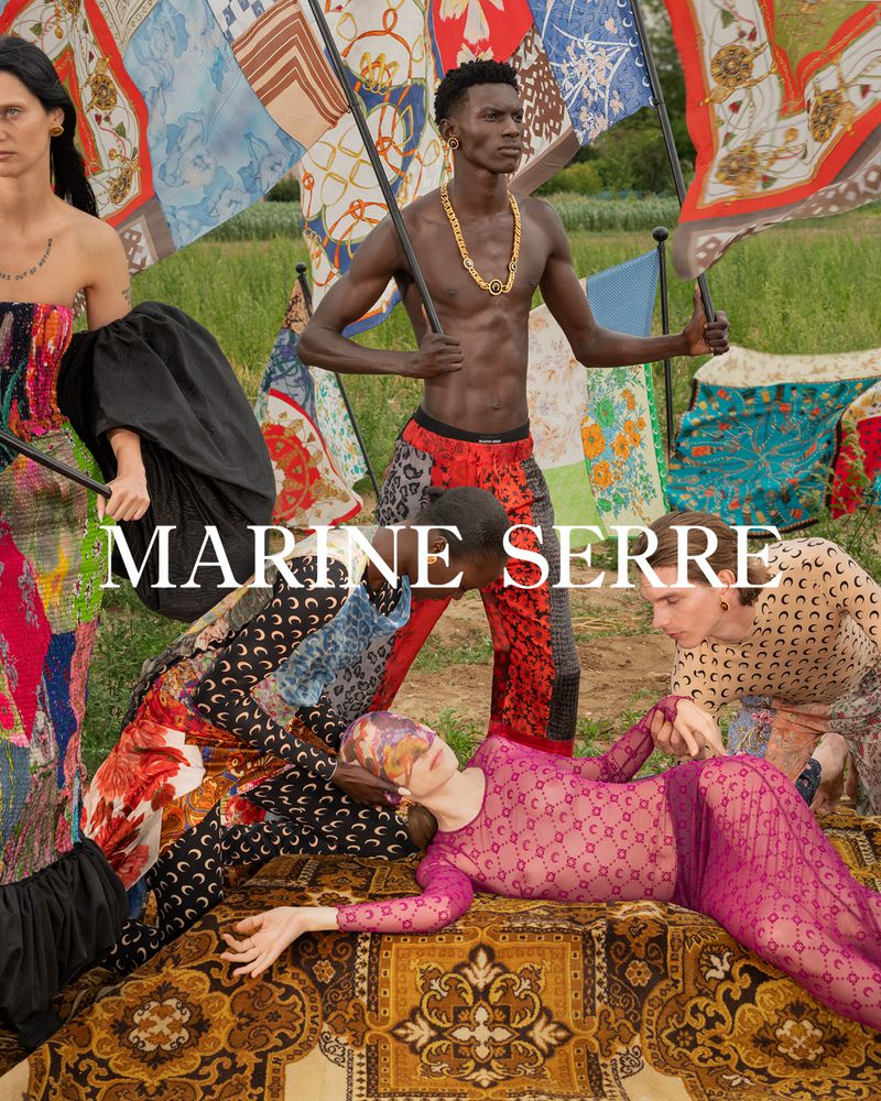 Editorial fashion image of a shirtless Black man holding flag poles, surrounded by a group of models in patterned clothing outdoors.
