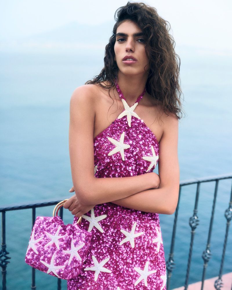 Adult female model wearing a pink halter dress with starfish standing in front of a seascape with a matching handbag.