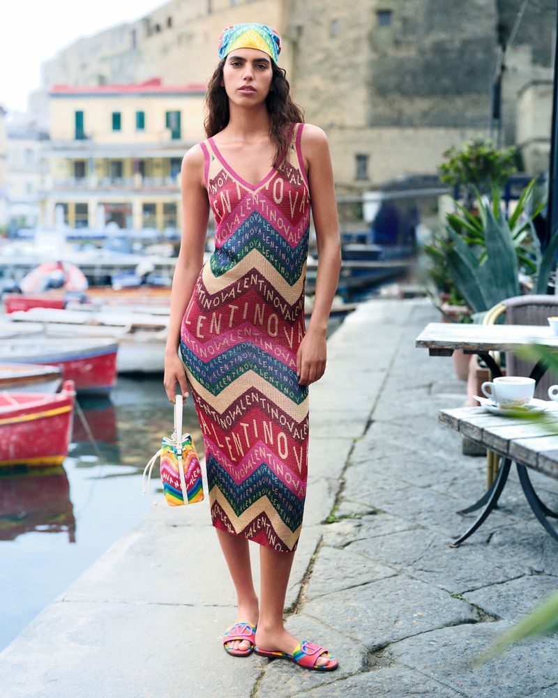 A woman poses on a stone pathway in a Valentino dress and matching headscarf, holding a rainbow bucket bag, in an urban harbor setting.