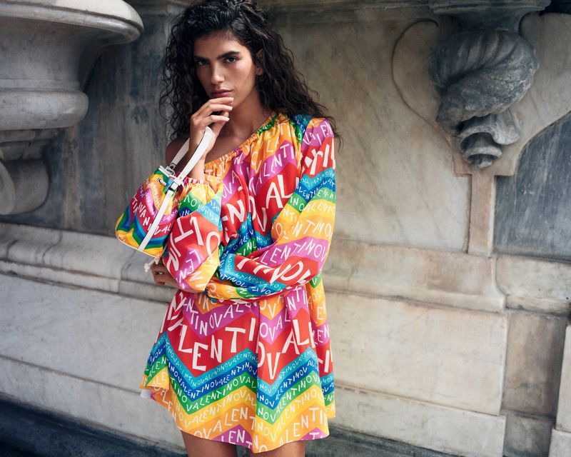 A woman poses in a colorful Valentino dress with a matching purse in front of a marble wall with decorative stonework.